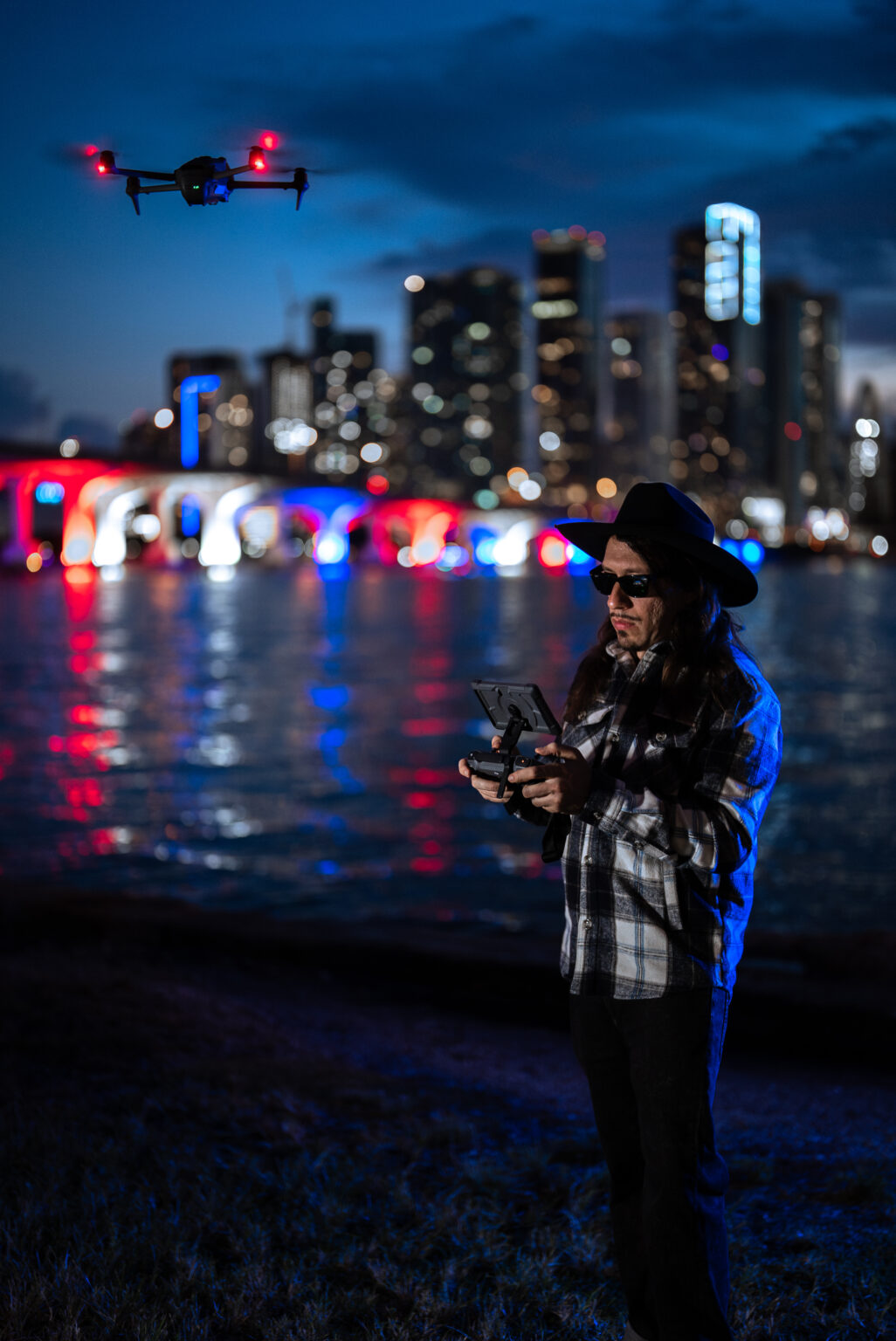 Matthew Hoyos flying his drone at night beside the water, with the lit-up bridge and Miami skyline behind him